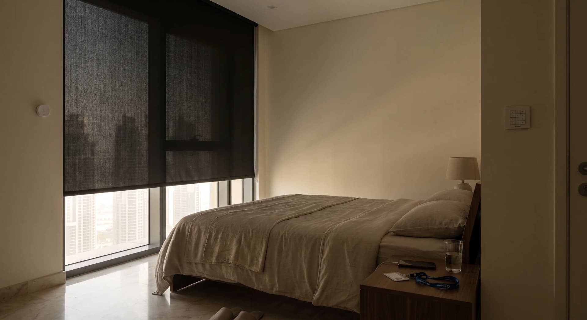 A dim, cool Dubai bedroom at mid-morning with a blackout roller blind almost fully down over a floor-to-ceiling window, a sliver of bright daylight and skyline at the bottom edge, a made bed with a folded work lanyard and a phone face-down on the nightstand, a small Aqara temperature sensor on the wall, and a low warm lamp switched off.