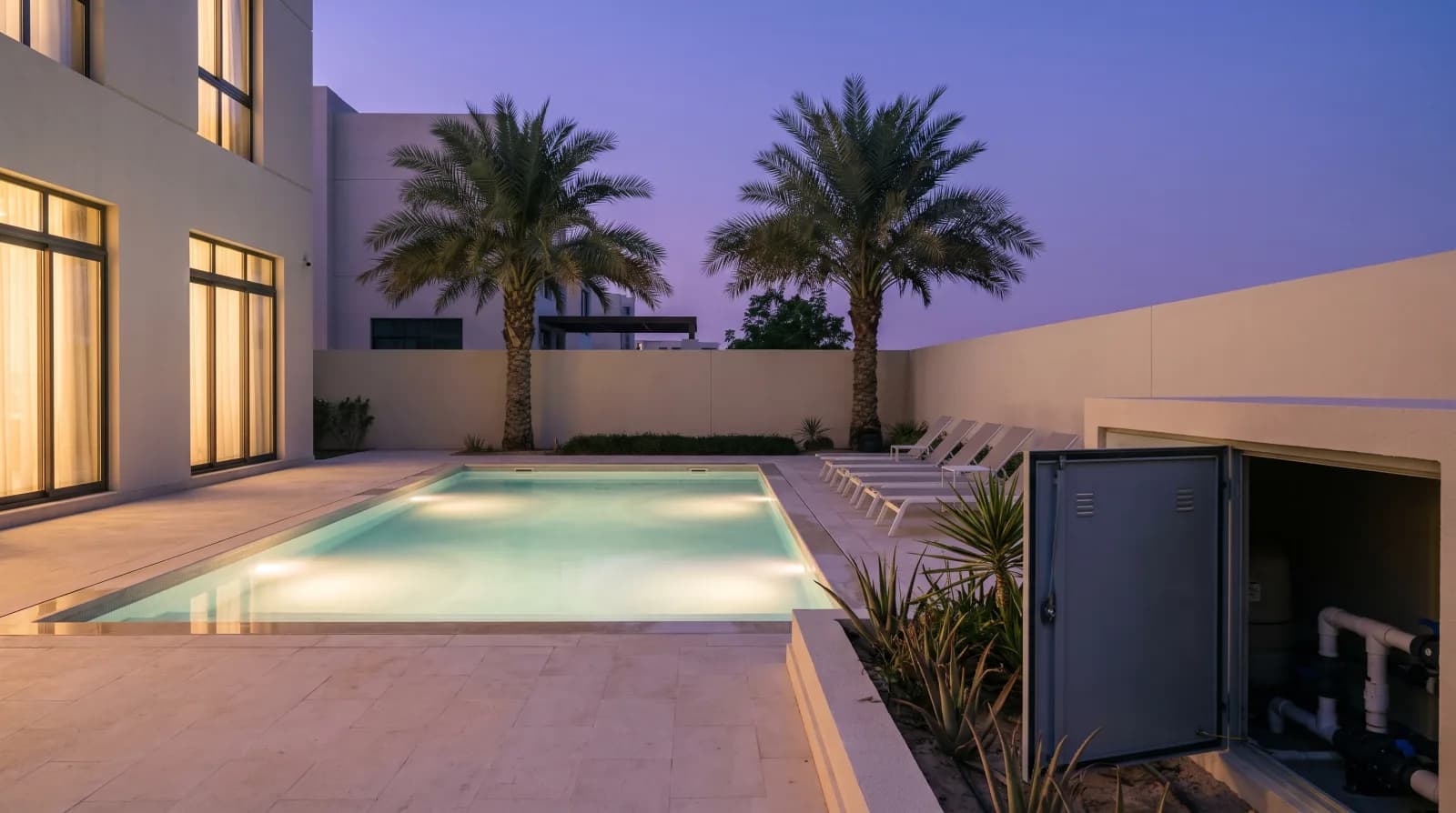 A Dubai villa garden at dusk with a lit swimming pool, two date palms and an open pump-room door showing the filter and pipework