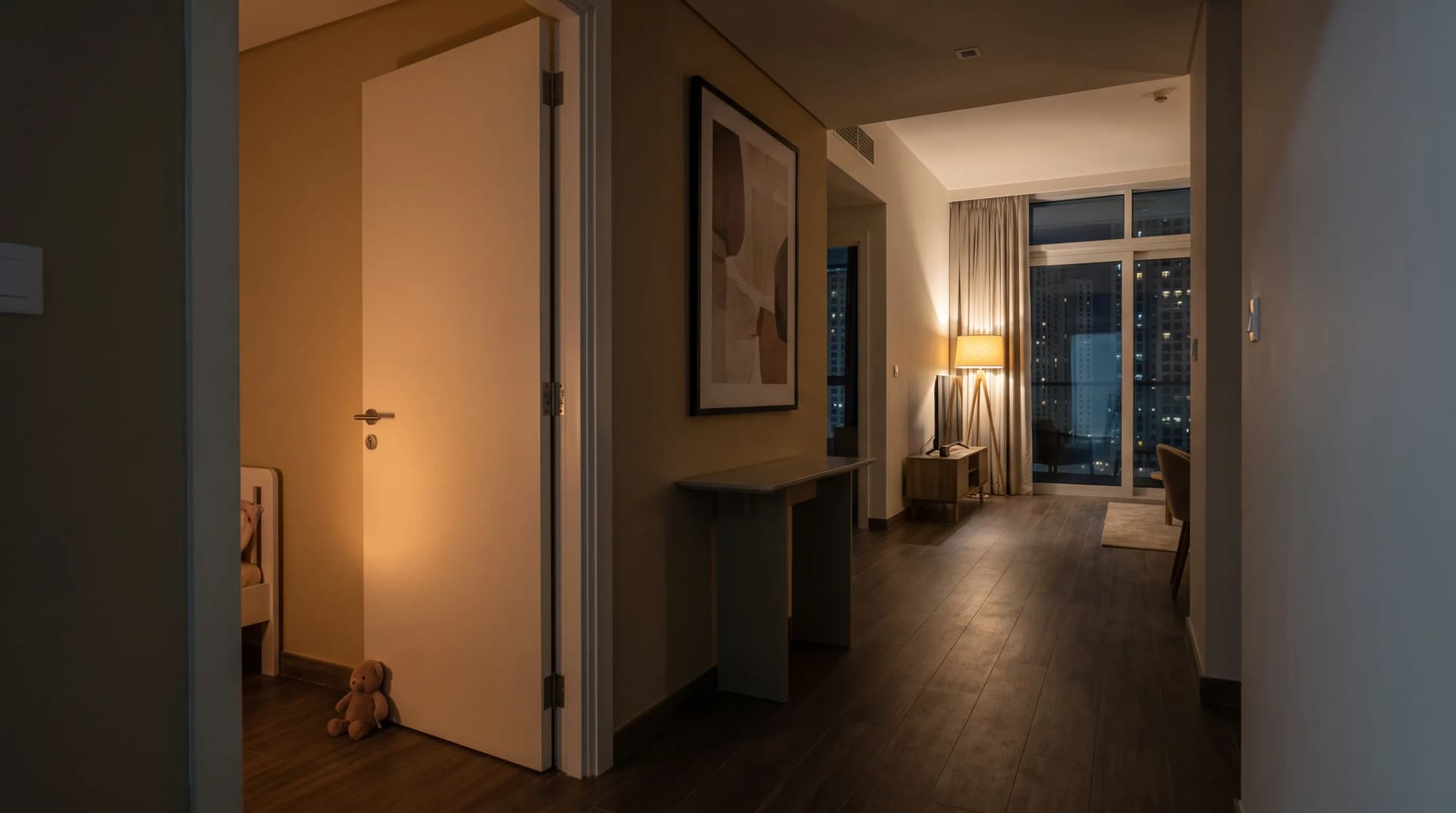 A Dubai apartment hallway at night, a child's bedroom door ajar with a teddy bear against it and warm light inside, and the living room beyond lit by a single lamp against the city skyline