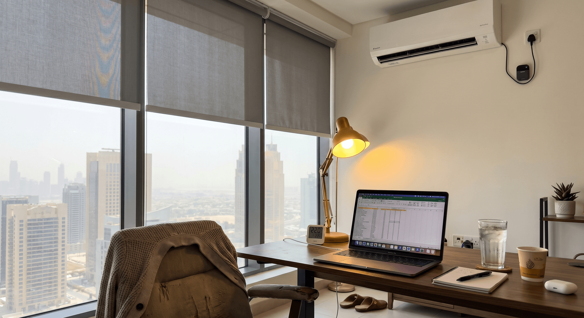 A Dubai apartment home office at 11am with floor-to-ceiling windows showing a bright hazy skyline, motorized roller blinds half-drawn to block glare, a Sensibo smart AC controller on the wall near a white split unit, an open MacBook on a walnut desk with a glass of water, Aqara temperature sensor on the shelf, warm desk lamp on, no people, lived-in