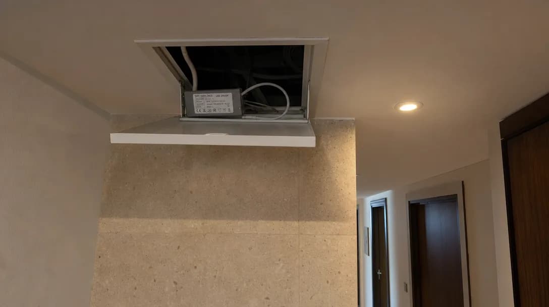 An open square gypsum ceiling access hatch in a Dubai apartment hallway, a small grey LED driver box sitting just inside the ceiling void with its printed specification label visible and white cabling coiled behind it, and a warm recessed downlight lit in the ceiling beside the hatch