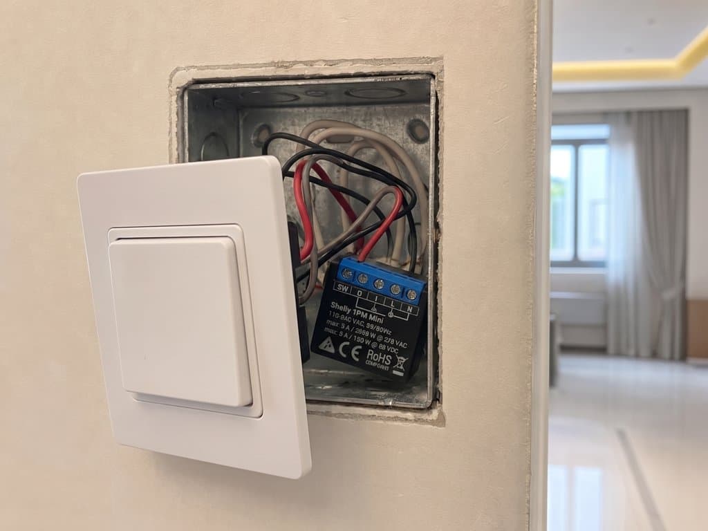 Open wall back-box showing a Shelly 1PM Mini relay tucked behind an existing brushed-nickel rocker switch in a Dubai townhouse